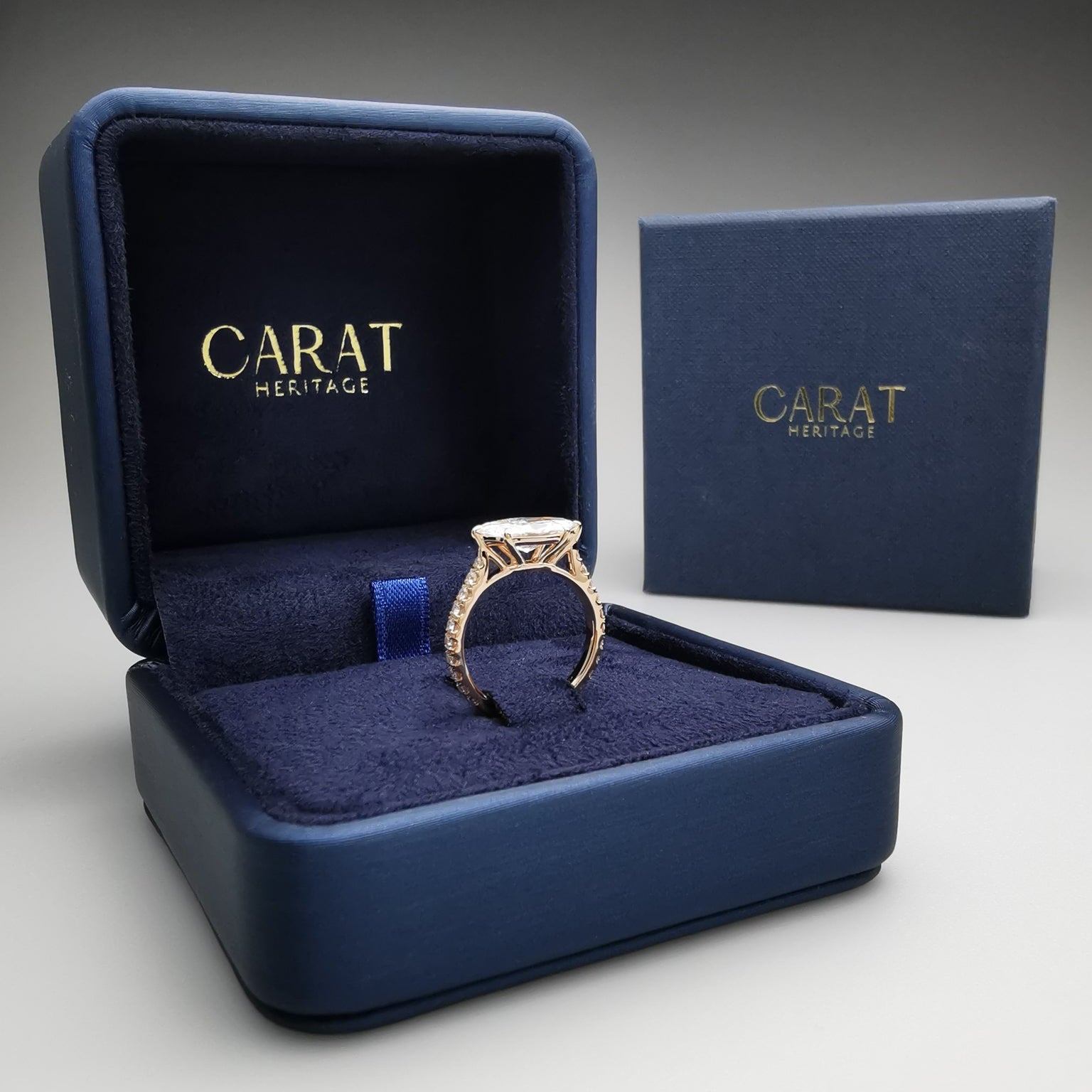 Gold ring in a blue 'Carat Heritage' jewelry box on a gray background