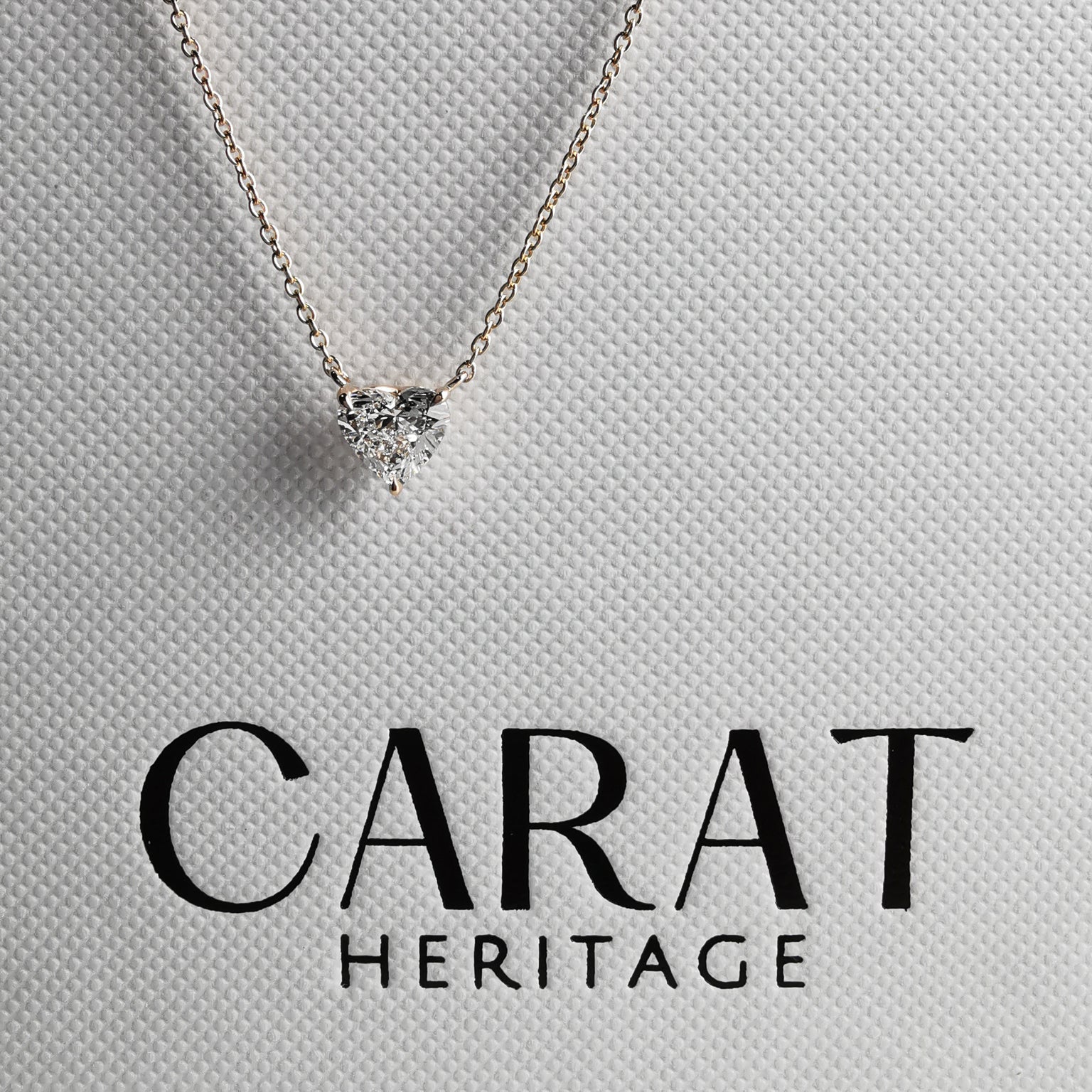Heart-shaped diamond necklace on a light gray background with 'Carat Heritage' branding.