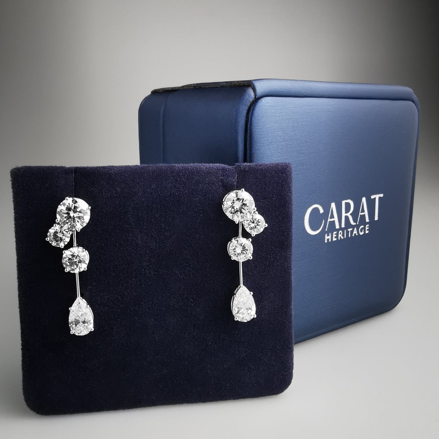 Diamond earrings on a black stand with a blue 'Carat Heritage' box in the background