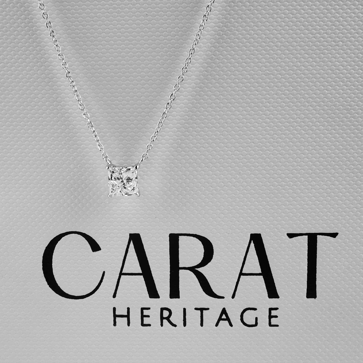 Silver necklace with a diamond pendant on a gray background, featuring the Carat Heritage logo.