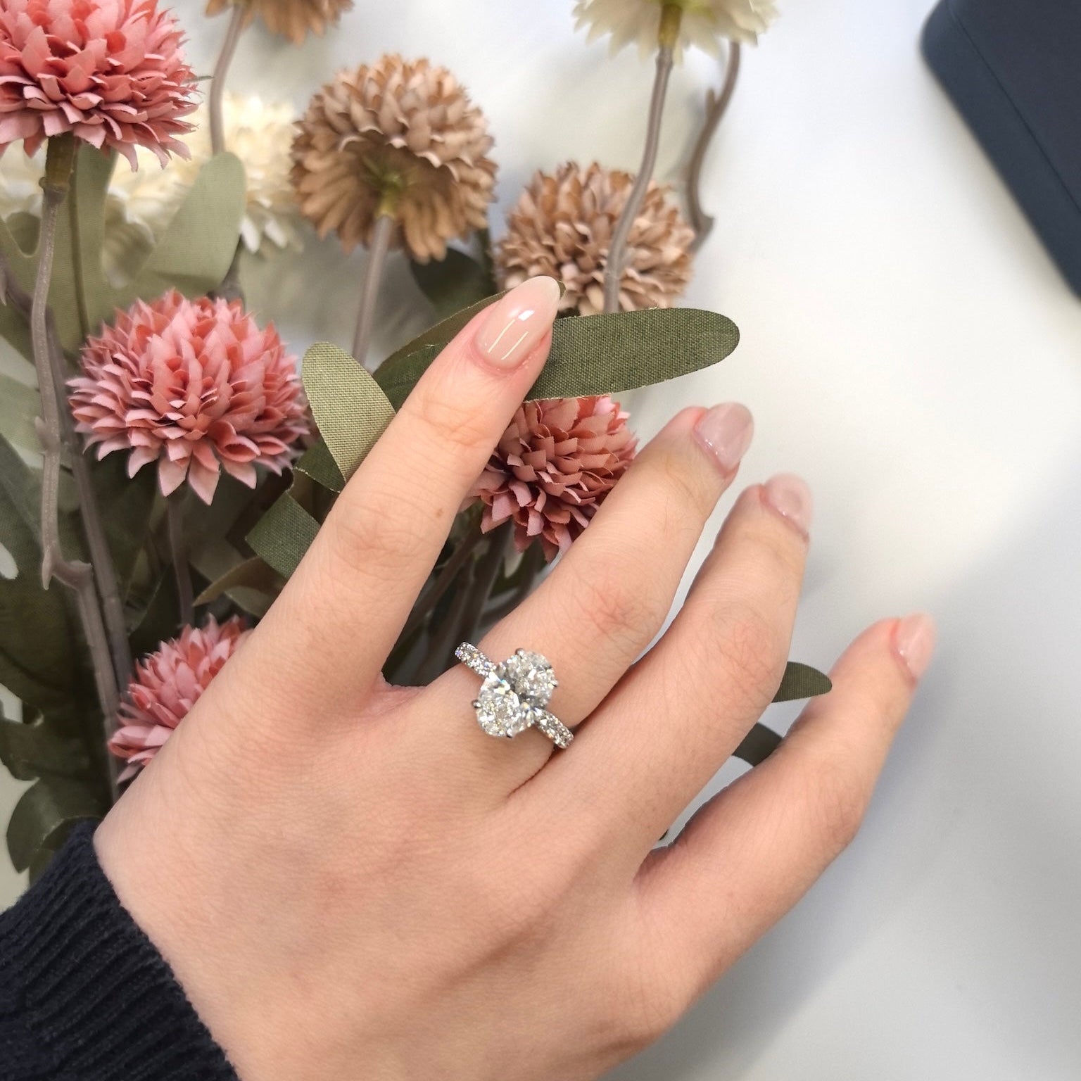 Hand wearing a oval diamond ring with flowers in the background